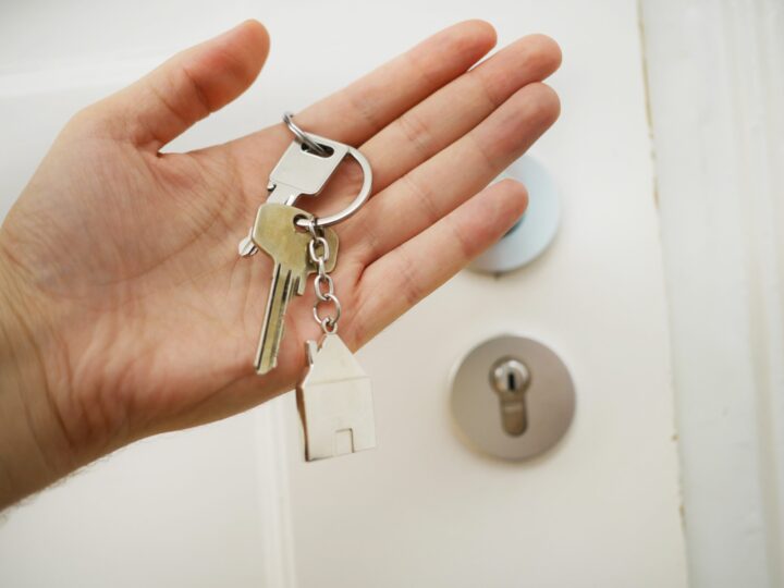 Hand holding a keychain with two shiny keys in front of a wooden door, symbolizing new homeownership.
