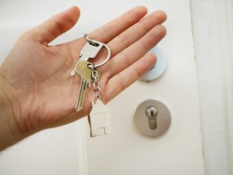 Hand holding a keychain with two shiny keys in front of a wooden door, symbolizing new homeownership.